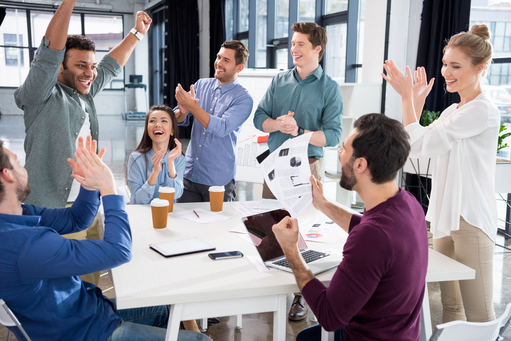 A sales team sitting around a table with one man raising his arms in celebration.